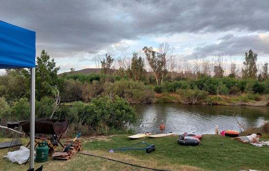 Grass terrace below campsite, with paddle boards, tubes, guy reading, and boy swimming in river.